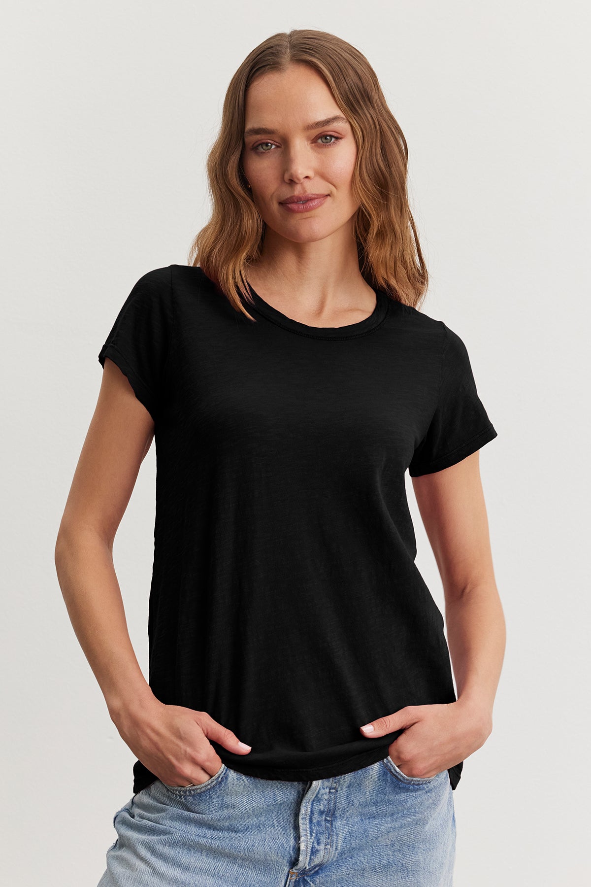   A person with wavy hair stands with hands in pockets against a plain background, wearing Velvet by Graham & Spencer's black TILLY TEE and jeans, embodying a true California classic style. 