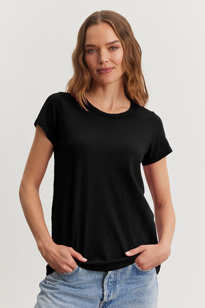 A person with wavy hair stands with hands in pockets against a plain background, wearing Velvet by Graham & Spencer's black TILLY TEE and jeans, embodying a true California classic style.