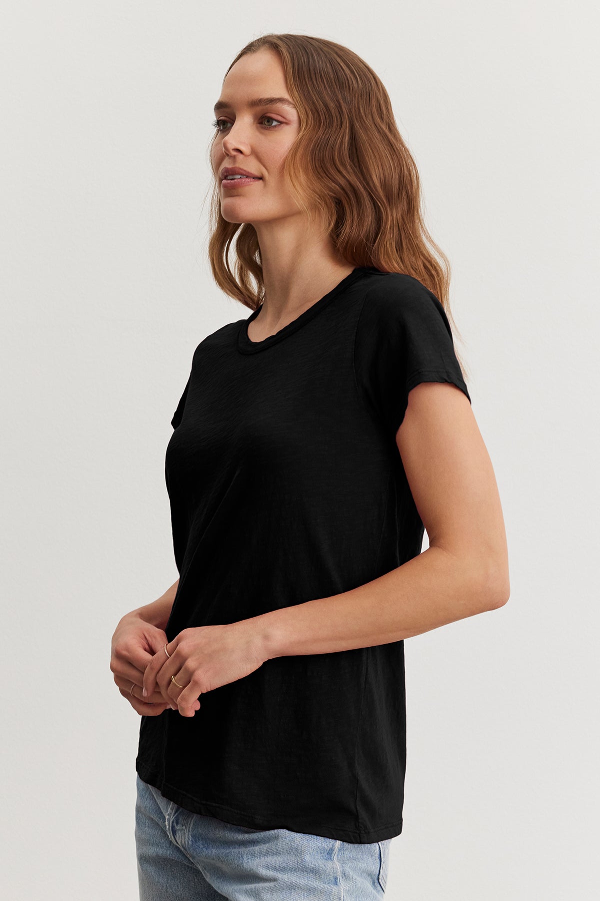   A woman with long wavy hair stands in a TILLY TEE by Velvet by Graham & Spencer and jeans, looking to her left against a plain background. 