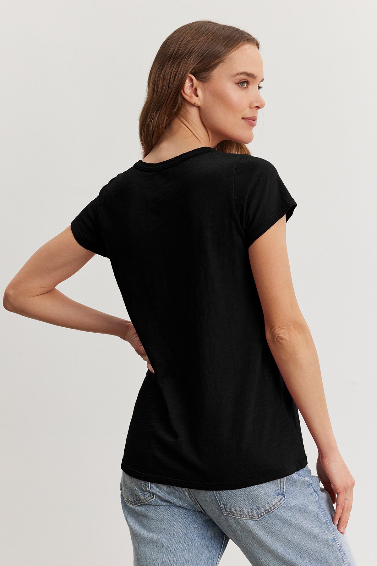 A woman with brown hair, wearing a TILLY TEE by Velvet by Graham & Spencer in black and jeans, stands sideways against a plain background.-38866044911809