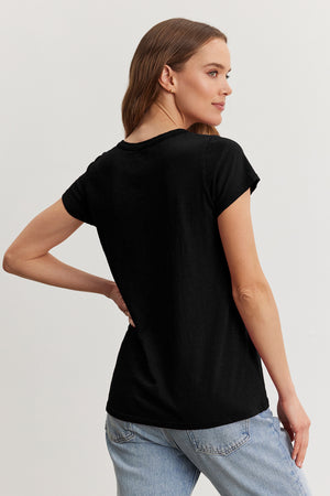 A woman with brown hair, wearing a TILLY TEE by Velvet by Graham & Spencer in black and jeans, stands sideways against a plain background.