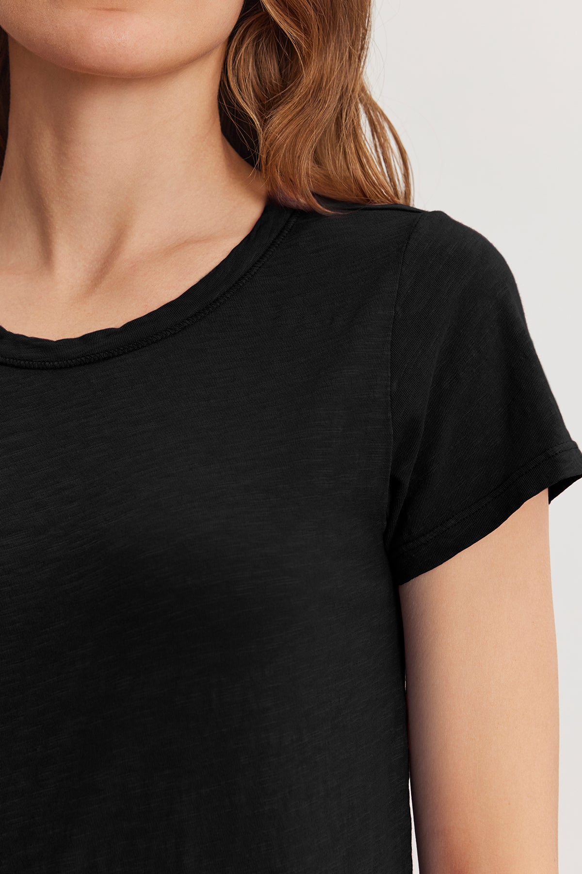   Someone with long hair is wearing the TILLY TEE from Velvet by Graham & Spencer, featuring a plain black crew neck design that embodies an effortless California classic style. 