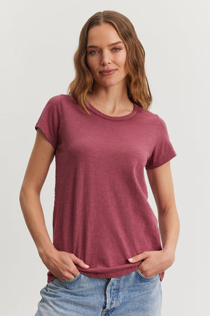 A person in the TILLY TEE by Velvet by Graham & Spencer and blue jeans poses with hands in pockets against a plain background.