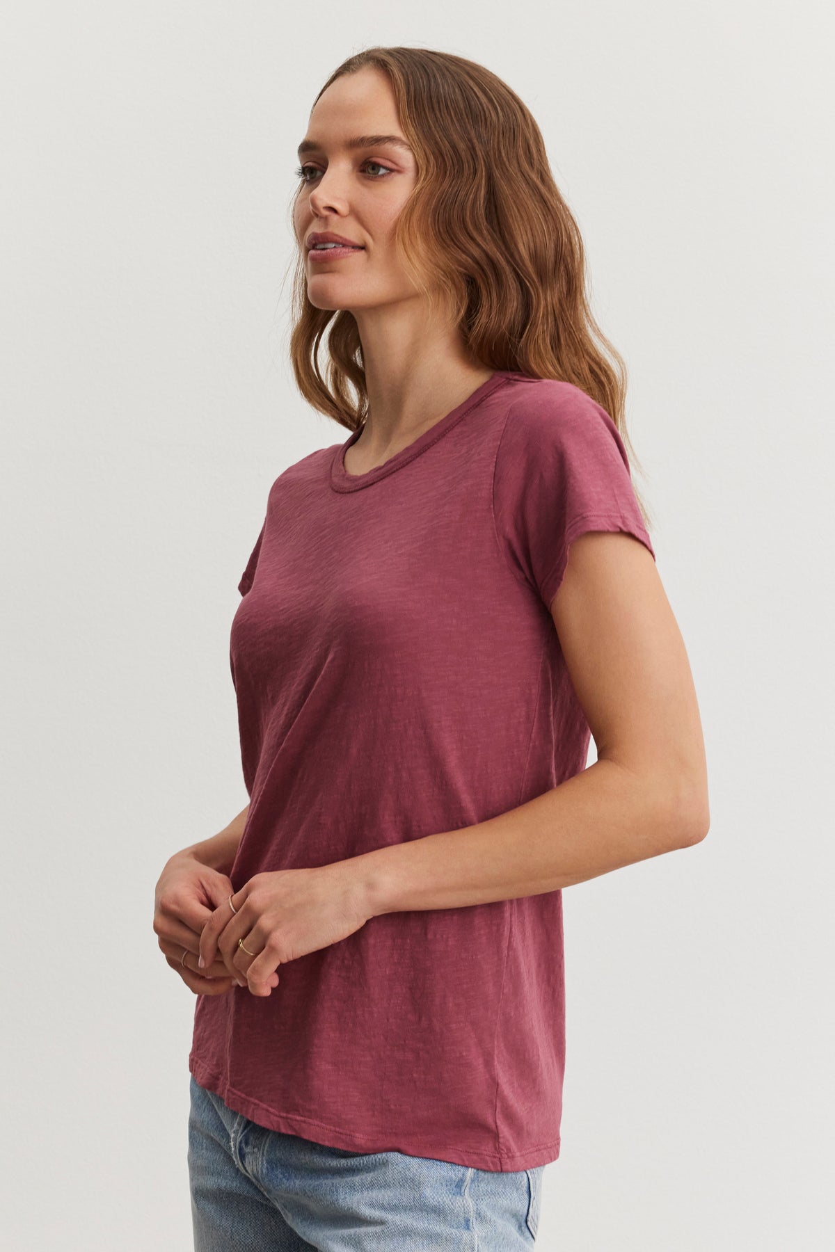   A person with long hair wears a TILLY TEE by Velvet by Graham & Spencer, a maroon soft cotton slub crew neck tee, paired with jeans, standing against a plain background. 