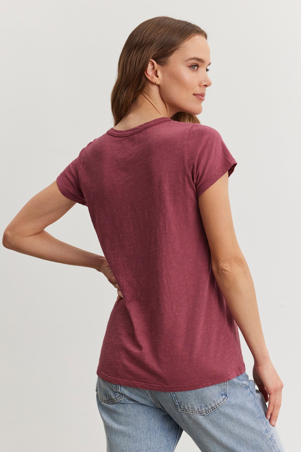   A person in a TILLY TEE by Velvet by Graham & Spencer, made from soft cotton slub in burgundy, paired with light blue jeans, stands sideways against a plain background. 