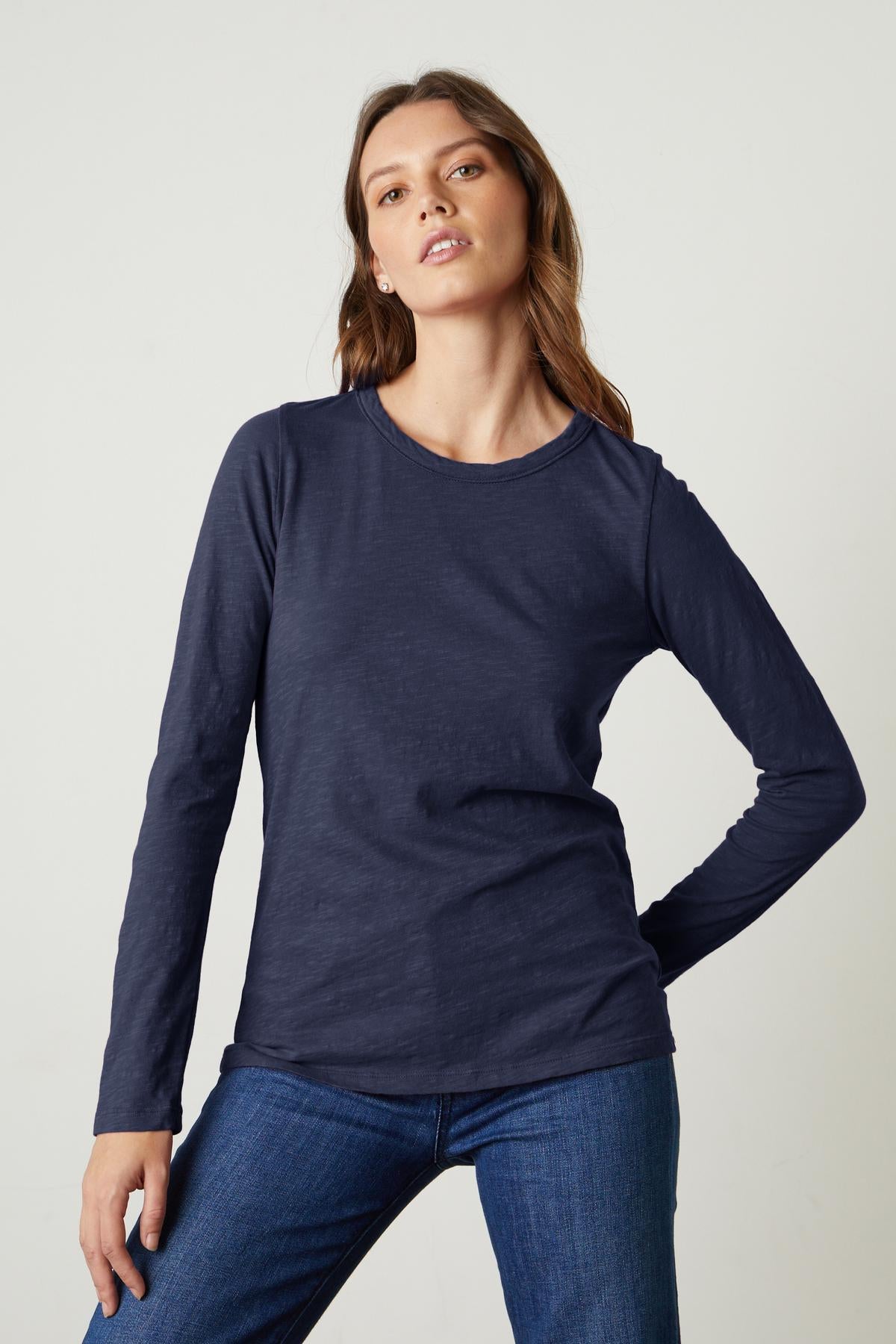   A person wearing a long-sleeved, navy blue LIZZIE TEE by Velvet by Graham & Spencer made of textured cotton slub with a classic crew neckline and jeans stands against a plain light background. 