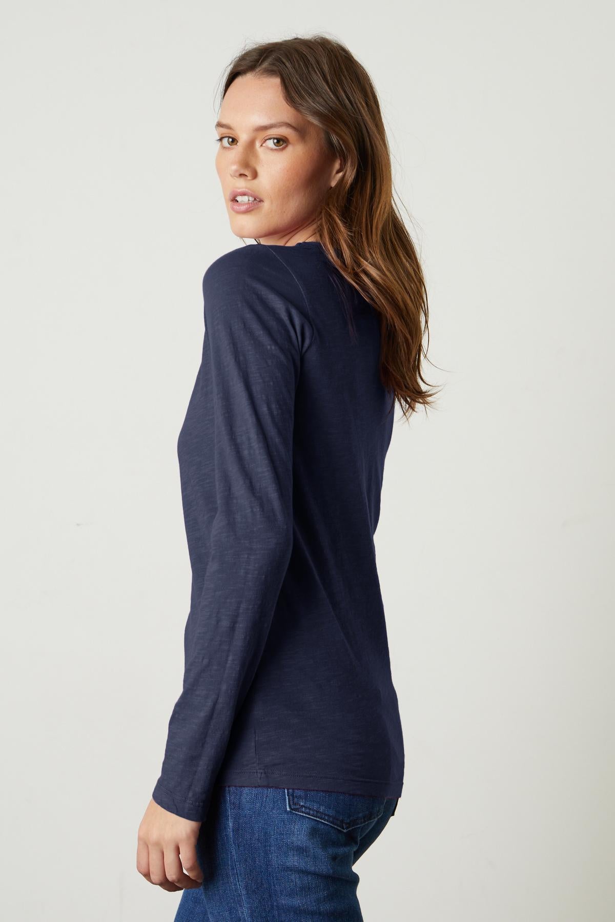   A woman with long brown hair, wearing a navy LIZZIE TEE from Velvet by Graham & Spencer and blue jeans, looks over her shoulder. 