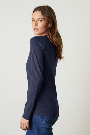 A woman with long brown hair, wearing a navy LIZZIE TEE from Velvet by Graham & Spencer and blue jeans, looks over her shoulder.