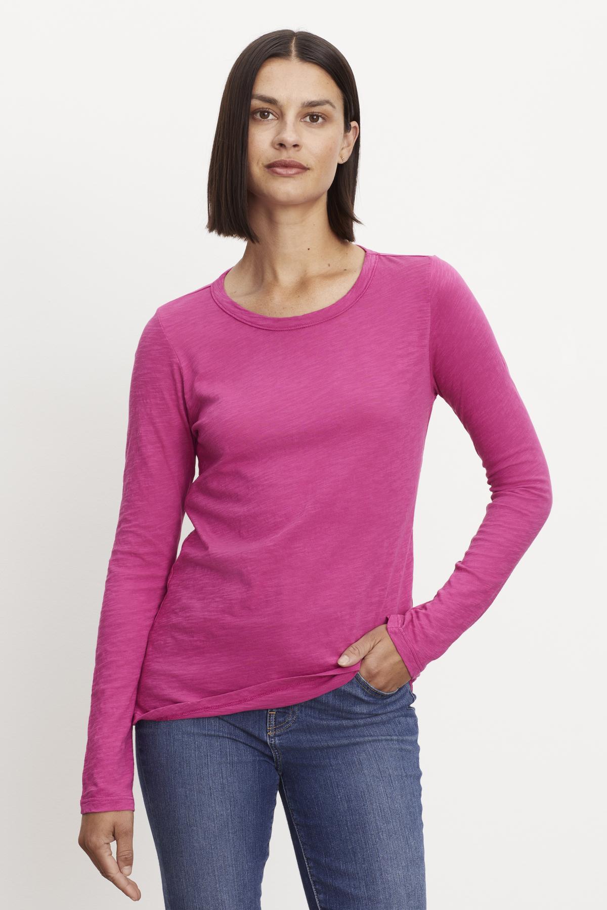   A woman with straight, shoulder-length hair, wearing a Velvet by Graham & Spencer LIZZIE TEE with a classic crew neckline and long sleeves paired with blue jeans, stands against a white background. She looks directly at the camera with a neutral expression. 