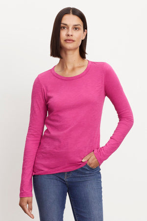 A woman with straight, shoulder-length hair, wearing a Velvet by Graham & Spencer LIZZIE TEE with a classic crew neckline and long sleeves paired with blue jeans, stands against a white background. She looks directly at the camera with a neutral expression.