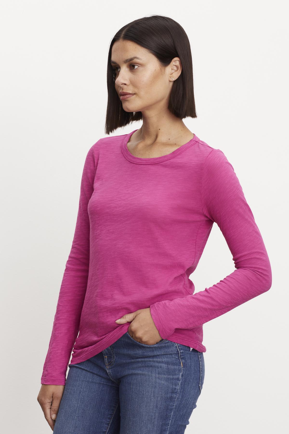   A woman with shoulder-length dark hair is wearing a Velvet by Graham & Spencer LIZZIE TEE, paired with blue jeans. She stands with one hand in her pocket against a plain white background. 