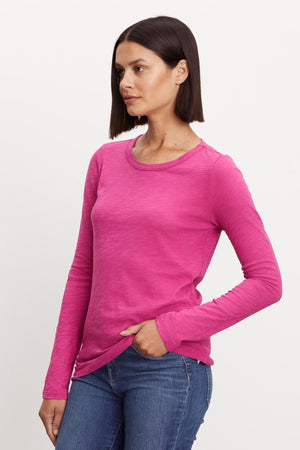 A woman with shoulder-length dark hair is wearing a Velvet by Graham & Spencer LIZZIE TEE, paired with blue jeans. She stands with one hand in her pocket against a plain white background.