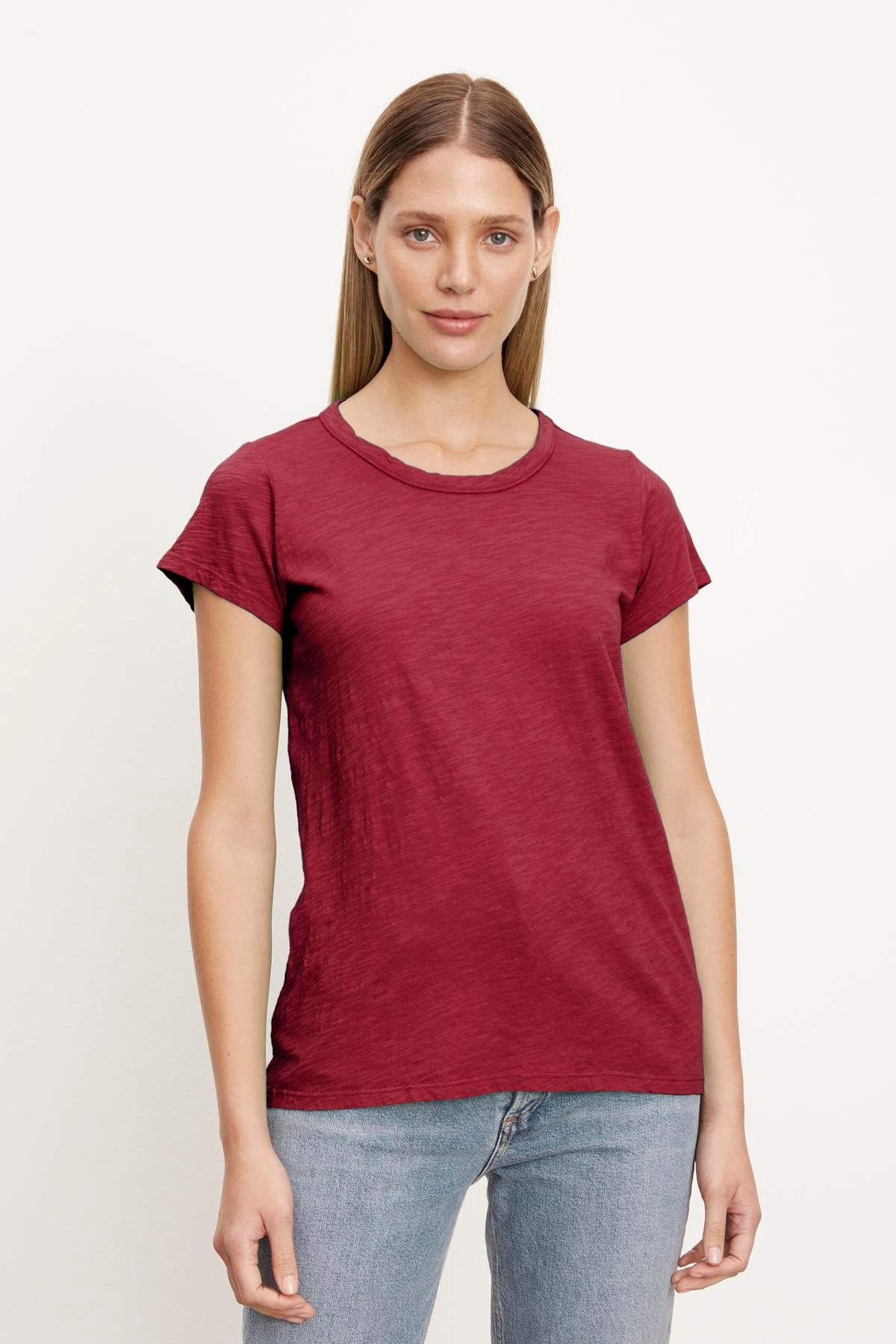   A person stands against a plain background, wearing a short-sleeved, red TILLY TEE by Velvet by Graham & Spencer and blue jeans, with a neutral expression. The perfect tee is made of soft cotton slub for ultimate comfort. 