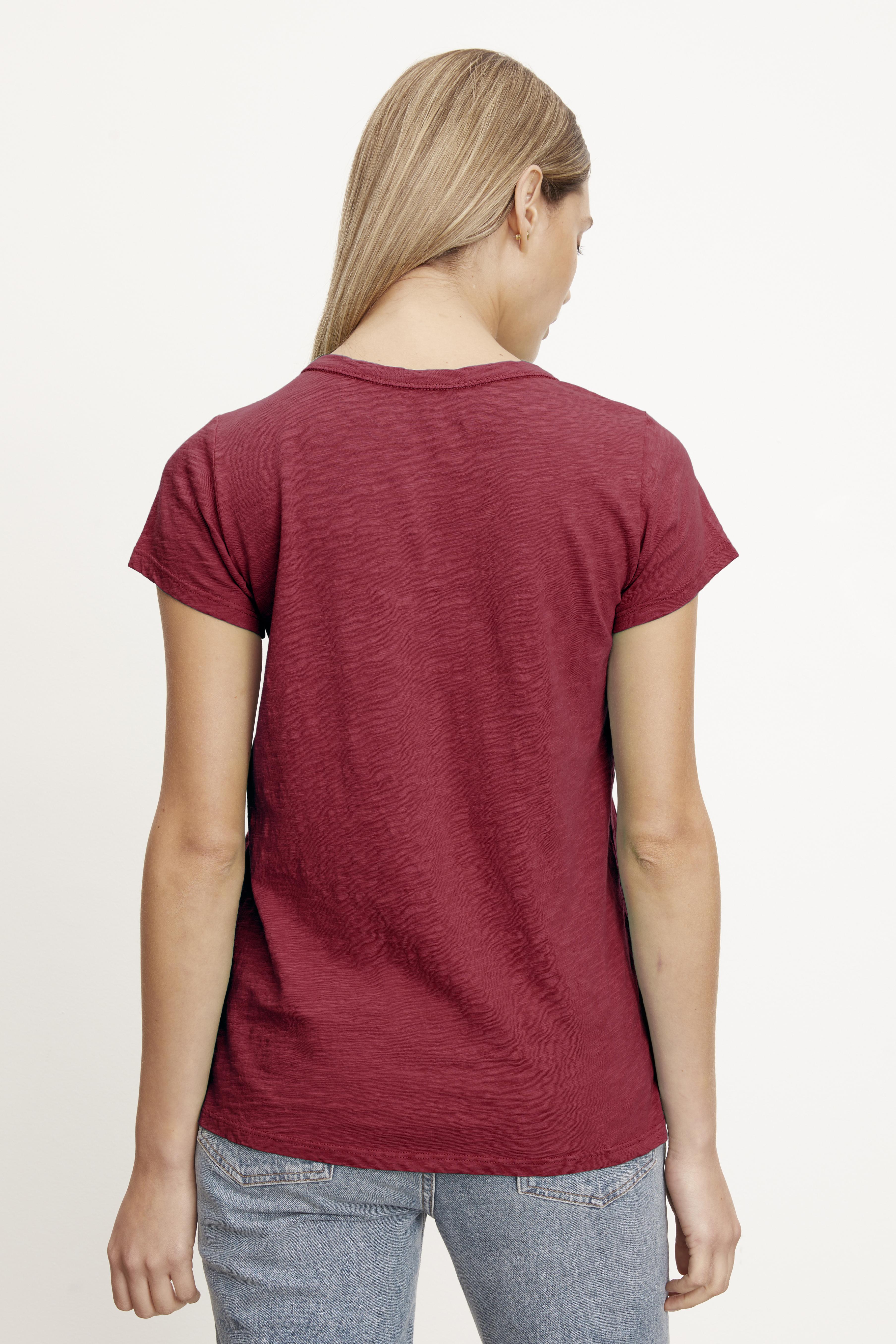   A person with long blonde hair, wearing a red TILLY TEE made by Velvet by Graham & Spencer and blue jeans, is standing with their back to the camera. 