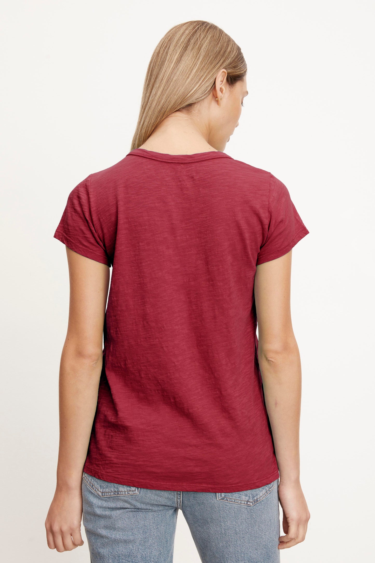 A person with long blonde hair, wearing a red TILLY TEE made by Velvet by Graham & Spencer and blue jeans, is standing with their back to the camera.-26883540353217