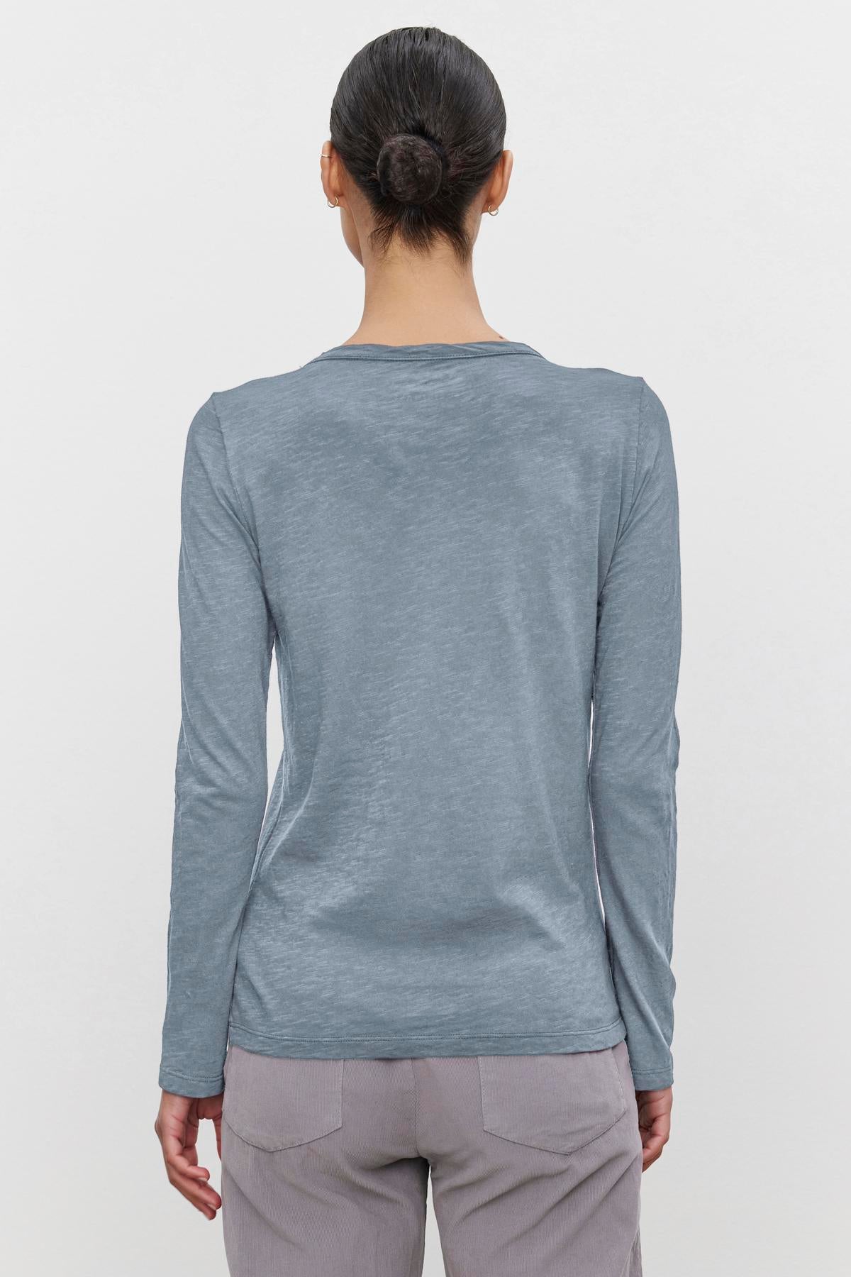   A person with hair in a bun is wearing the LIZZIE TEE by Velvet by Graham & Spencer, a long-sleeved, light blue textured cotton slub top, along with gray pants, and is facing away from the camera. 