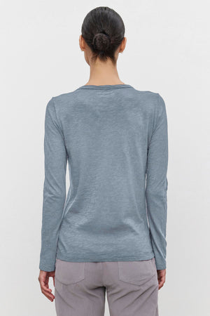 A person with hair in a bun is wearing the LIZZIE TEE by Velvet by Graham & Spencer, a long-sleeved, light blue textured cotton slub top, along with gray pants, and is facing away from the camera.