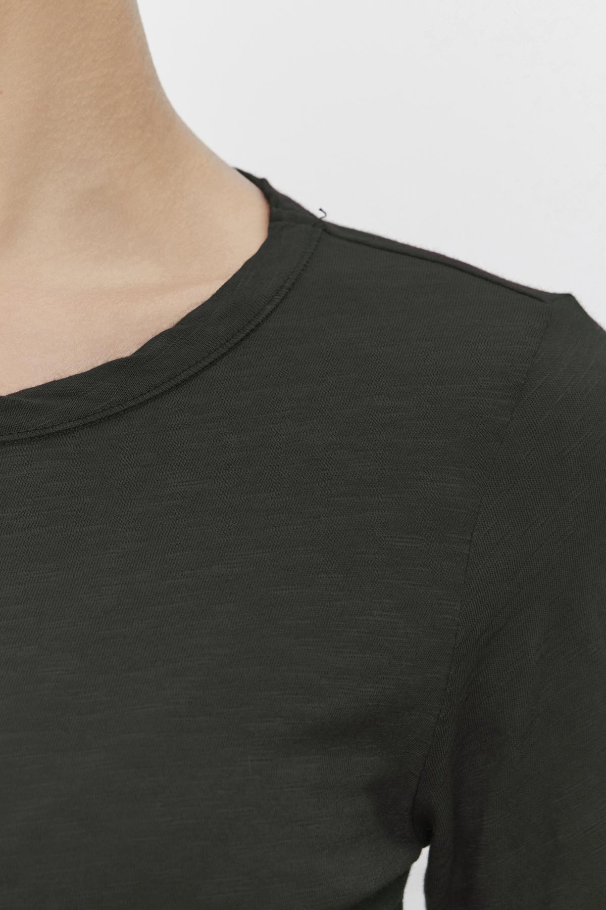   Close-up of a person wearing the LIZZIE TEE by Velvet by Graham & Spencer, featuring a classic crew neckline in dark green. The image highlights the shoulder and part of the upper chest against a neutral background, showcasing the depth and character added by its textured cotton slub fabric to this simple yet stylish piece. 