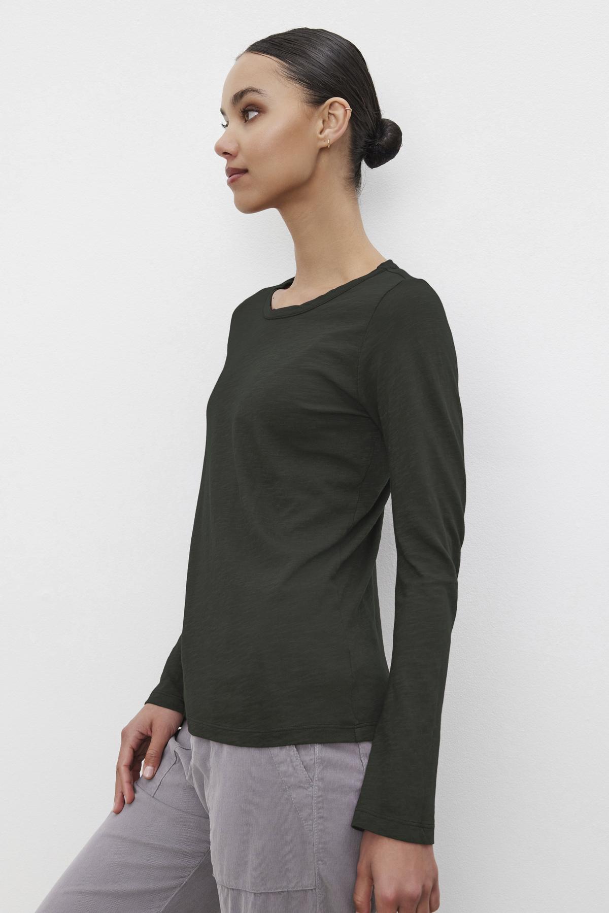   A woman wearing the Velvet by Graham & Spencer LIZZIE TEE, a textured cotton slub shirt in dark green, paired with light gray pants, stands against a white background, looking to the side. 