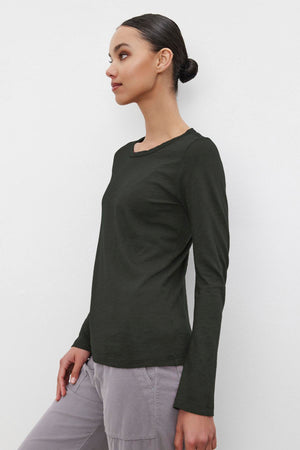 A woman wearing the Velvet by Graham & Spencer LIZZIE TEE, a textured cotton slub shirt in dark green, paired with light gray pants, stands against a white background, looking to the side.