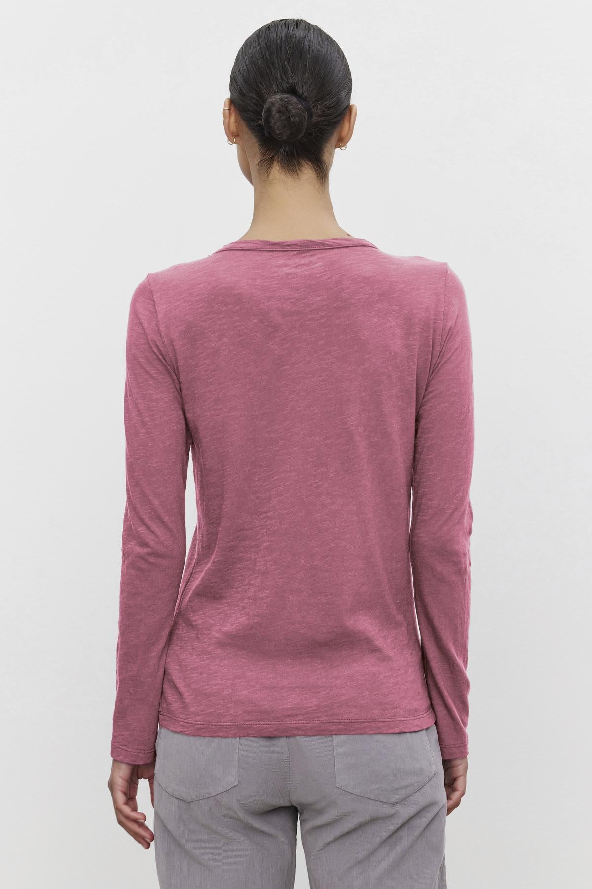   A person with a bun wears the Velvet by Graham & Spencer LIZZIE TEE, a long-sleeved pink shirt with a classic crew neckline, paired with gray pants, shown from the back against a plain white background. 