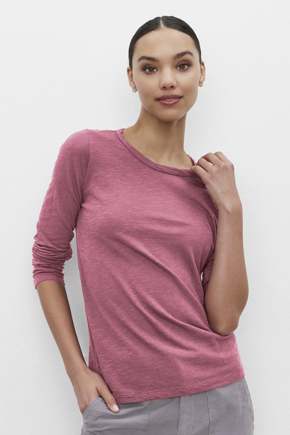 Someone wearing the Velvet by Graham & Spencer LIZZIE TEE, a long-sleeve pink top with a classic crew neckline, and gray pants stands against a plain white background, holding the neckline with one hand and resting the other in a pocket.-38642774802625