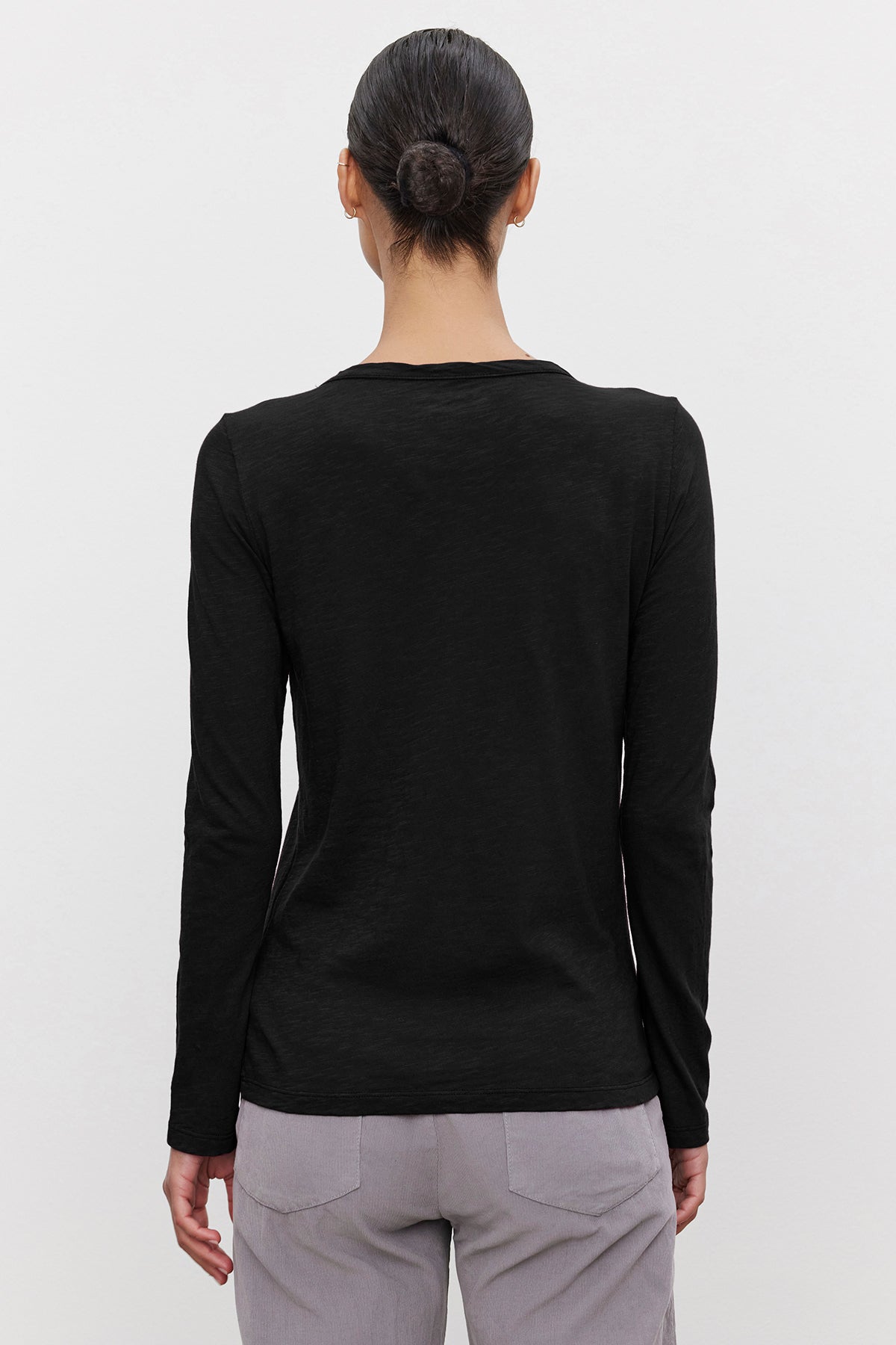   Back view of a person with tied-up hair, wearing a black long-sleeve LIZZIE TEE by Velvet by Graham & Spencer with a classic crew neckline and light gray pants, standing against a plain white background. 