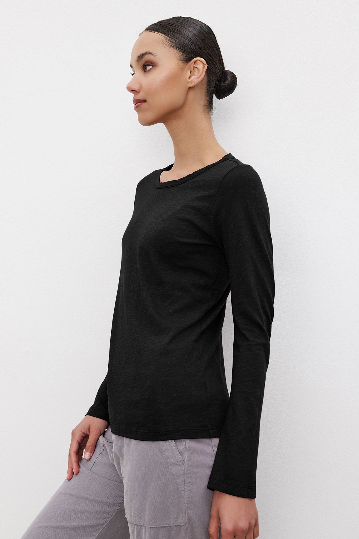   A woman with a sleek bun hairstyle is wearing the LIZZIE TEE by Velvet by Graham & Spencer, a long-sleeved black top made of textured cotton slub, and light gray pants. She is standing in profile against a plain white background. 