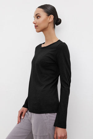 A woman with a sleek bun hairstyle is wearing the LIZZIE TEE by Velvet by Graham & Spencer, a long-sleeved black top made of textured cotton slub, and light gray pants. She is standing in profile against a plain white background.