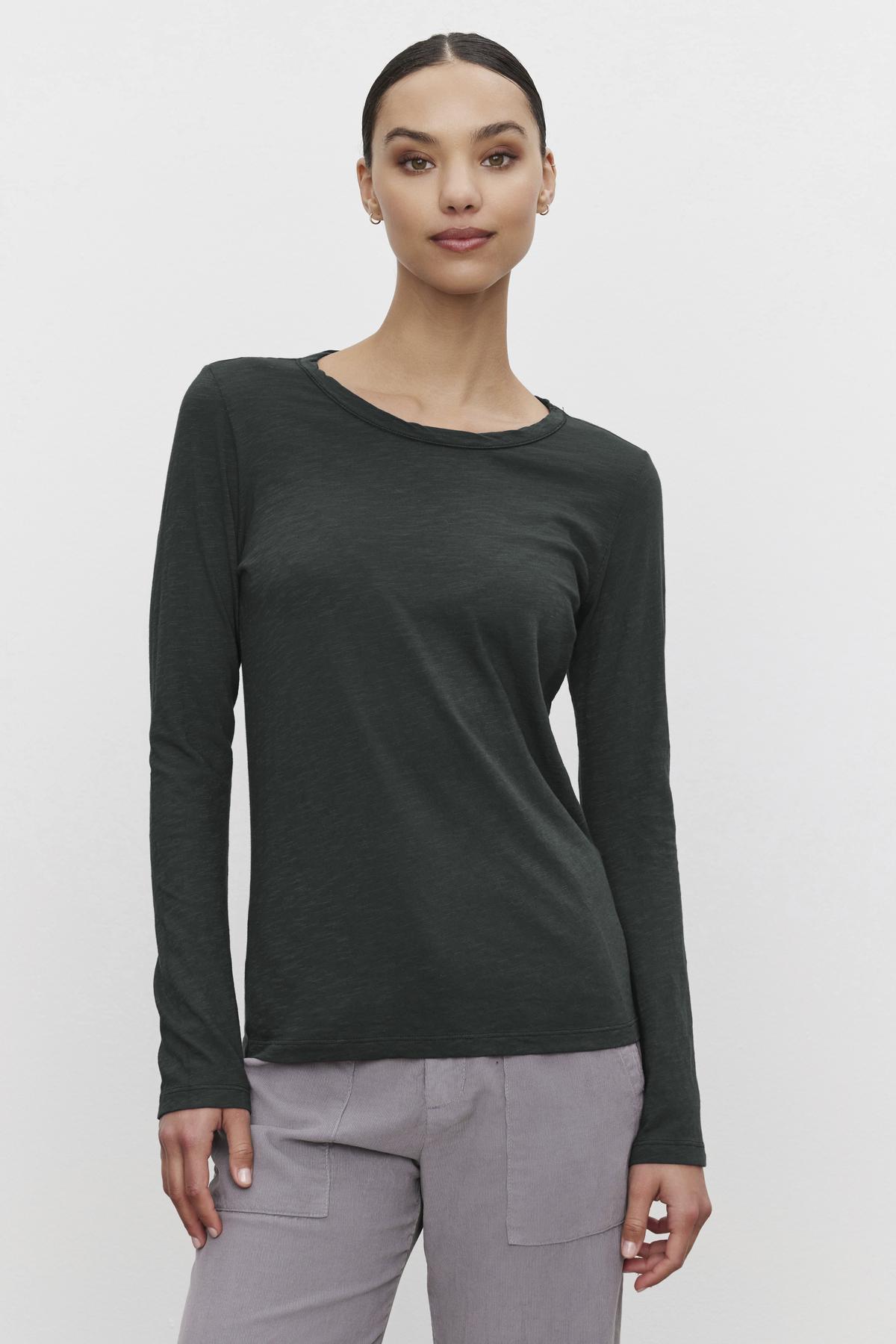 A person wearing a LIZZIE TEE by Velvet by Graham & Spencer, styled in dark green with a crew neckline and long sleeves, along with gray pants, stands against a plain background.-38139494301889