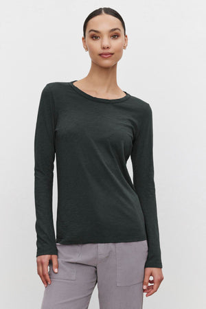 A person wearing a LIZZIE TEE by Velvet by Graham & Spencer, styled in dark green with a crew neckline and long sleeves, along with gray pants, stands against a plain background.