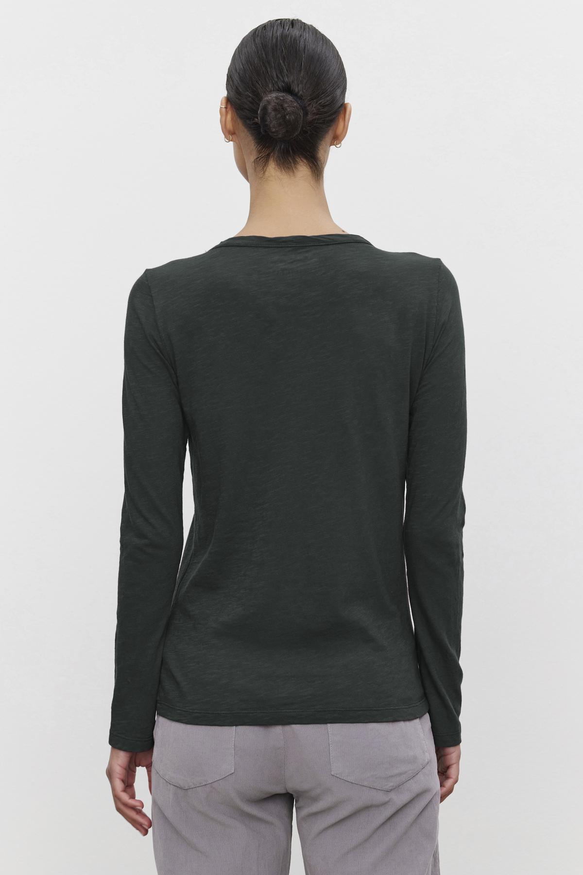   A person with dark hair in a bun wears the LIZZIE TEE by Velvet by Graham & Spencer, a long-sleeved dark green shirt crafted from cotton slub, along with light gray pants, facing away. 