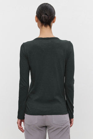 A person with dark hair in a bun wears the LIZZIE TEE by Velvet by Graham & Spencer, a long-sleeved dark green shirt crafted from cotton slub, along with light gray pants, facing away.