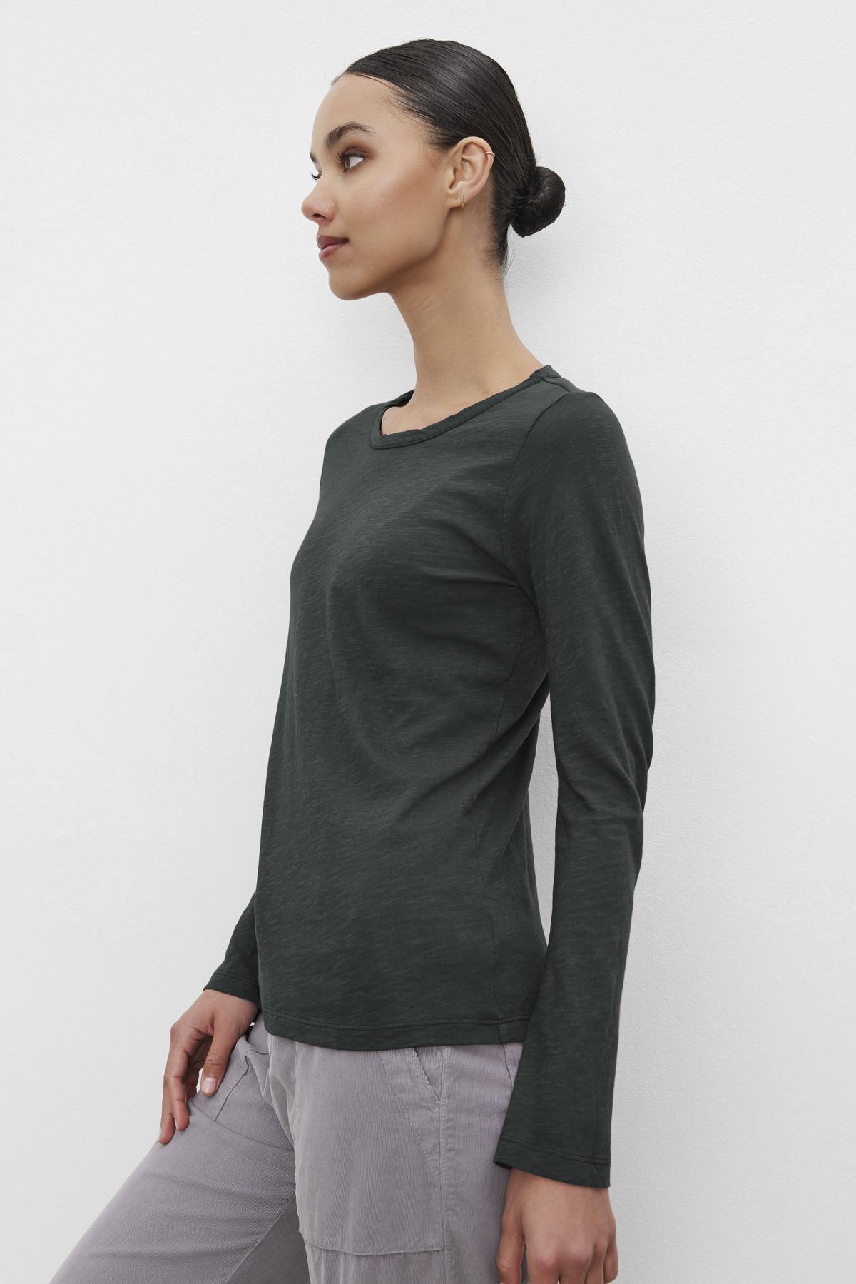   Someone wearing the LIZZIE TEE, a dark green long-sleeve shirt with a crew neckline from Velvet by Graham & Spencer, stands against a plain white background. 