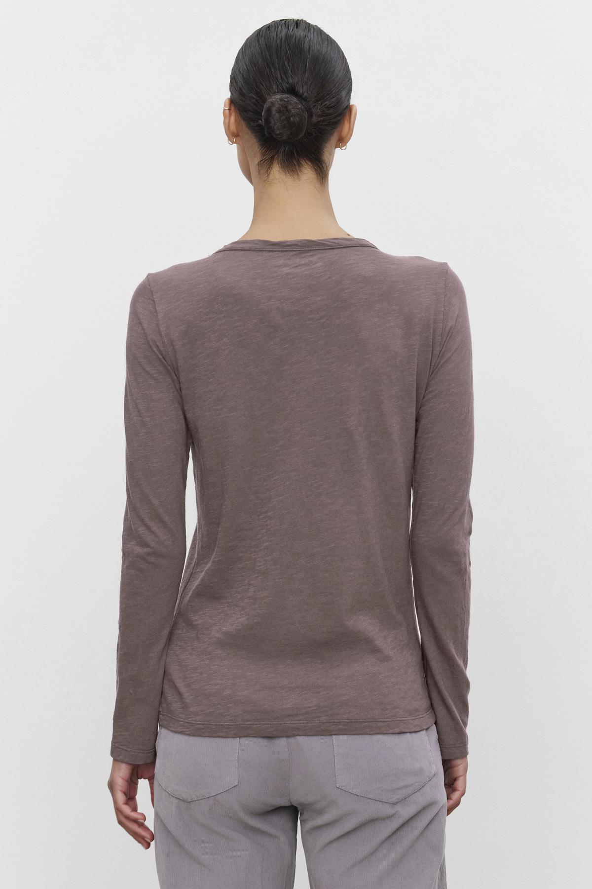   A person with dark hair in a bun is wearing the LIZZIE TEE from Velvet by Graham & Spencer, a long-sleeved brown shirt with a classic crew neckline, paired with light-colored pants, standing with their back to the camera against a plain white background. 