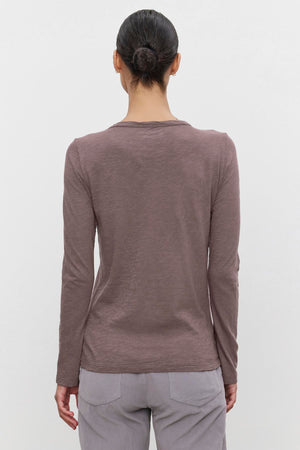 A person with dark hair in a bun is wearing the LIZZIE TEE from Velvet by Graham & Spencer, a long-sleeved brown shirt with a classic crew neckline, paired with light-colored pants, standing with their back to the camera against a plain white background.