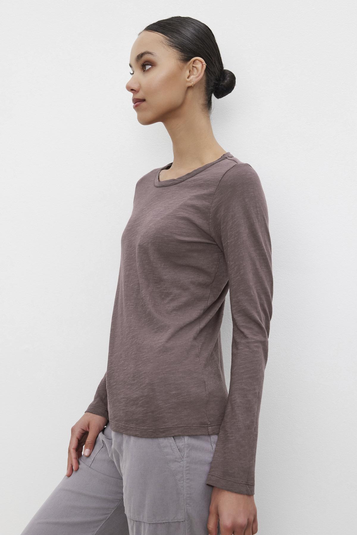   A person with hair styled in a bun stands against a white background, wearing the Long-Sleeve LIZZIE TEE by Velvet by Graham & Spencer, featuring a classic crew neckline, paired with gray pants. 