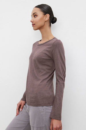 A person with hair styled in a bun stands against a white background, wearing the Long-Sleeve LIZZIE TEE by Velvet by Graham & Spencer, featuring a classic crew neckline, paired with gray pants.