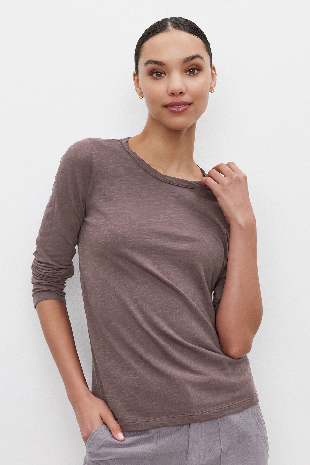 Person wearing the LIZZIE TEE, a long-sleeve, brown shirt by Velvet by Graham & Spencer, featuring a classic crew neckline. One hand touches the collar while the other rests in a pocket as they stand against a plain white background.-37935126020289