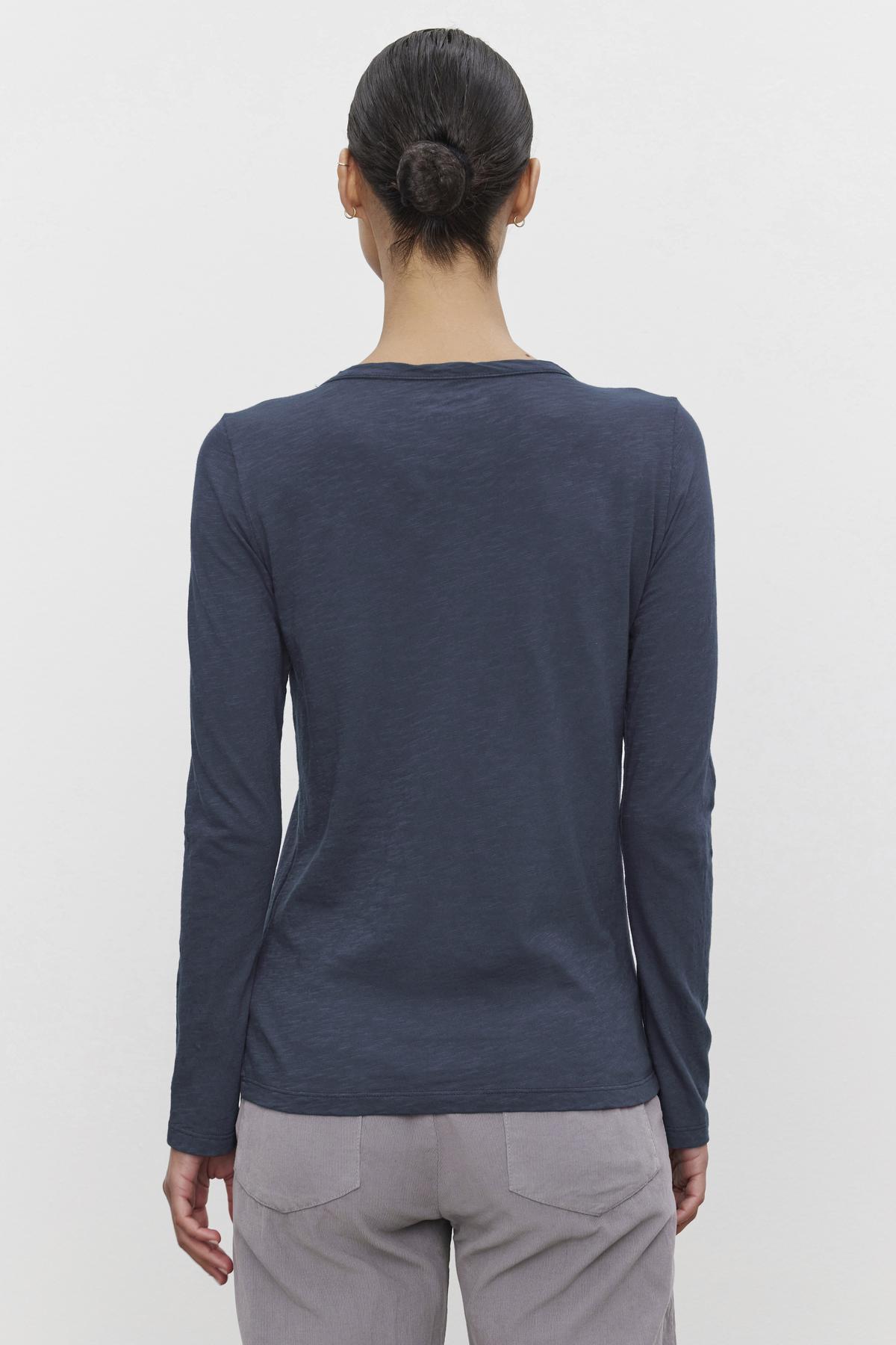 A person with dark hair in a bun is shown from the back, wearing the Velvet by Graham & Spencer LIZZIE TEE, a long-sleeve, dark blue shirt with a classic crew neckline, paired with light-colored pants.-37935125856449