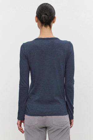 A person with dark hair in a bun is shown from the back, wearing the Velvet by Graham & Spencer LIZZIE TEE, a long-sleeve, dark blue shirt with a classic crew neckline, paired with light-colored pants.