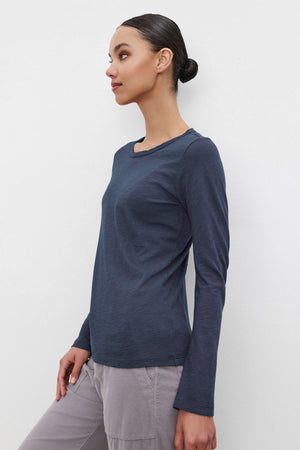 A person with their hair in a bun wears the Velvet by Graham & Spencer LIZZIE TEE, a long-sleeved, navy blue shirt with a classic crew neckline, paired with light-colored pants. They are turned slightly to the side against a plain white background.