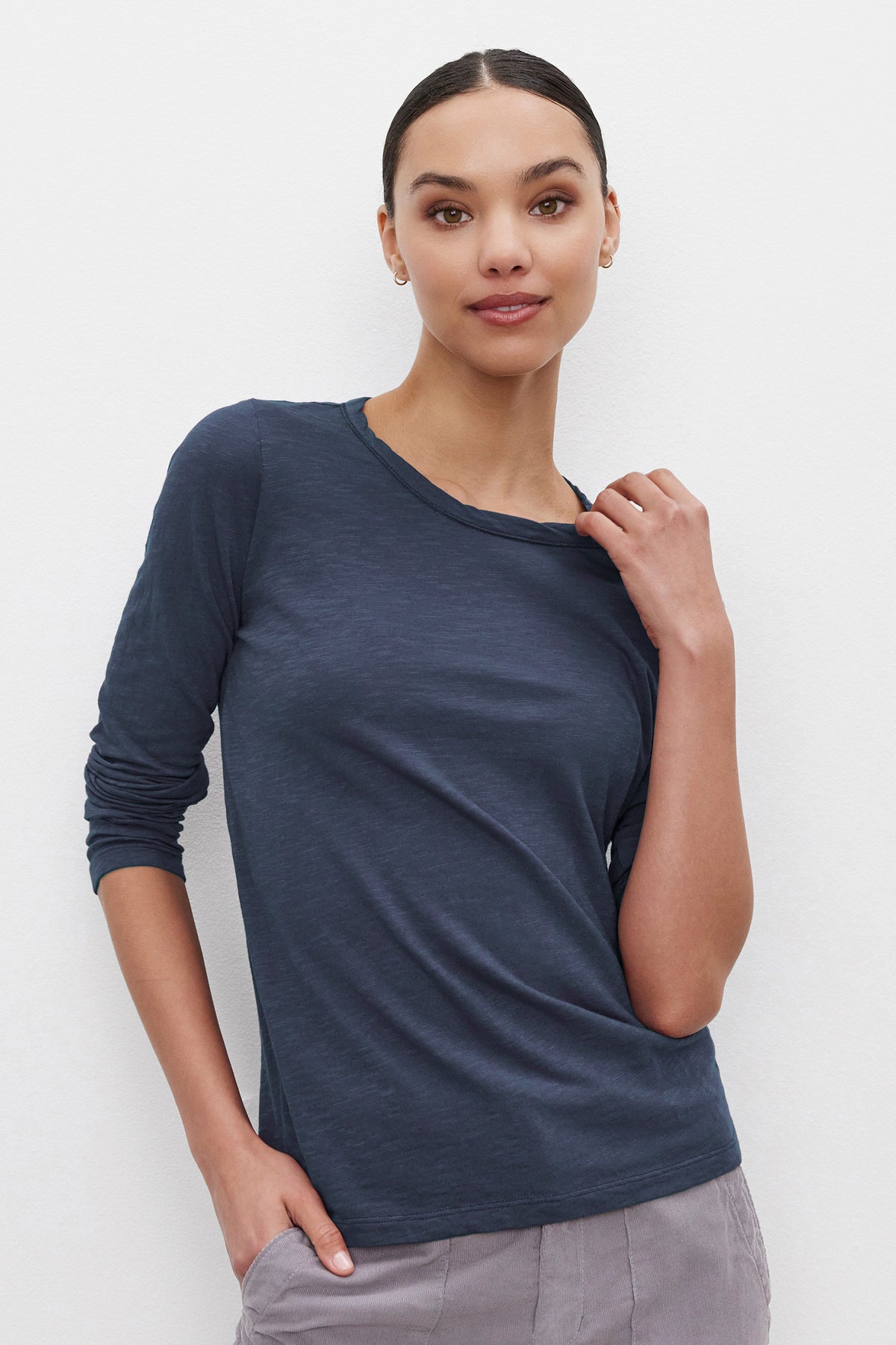   A person with dark, pulled-back hair, wearing the LIZZIE TEE from Velvet by Graham & Spencer – a classic crew neckline long-sleeve top in dark blue – paired with light-colored pants, stands against a light background, touching their collar with one hand and placing the other in their pocket. 