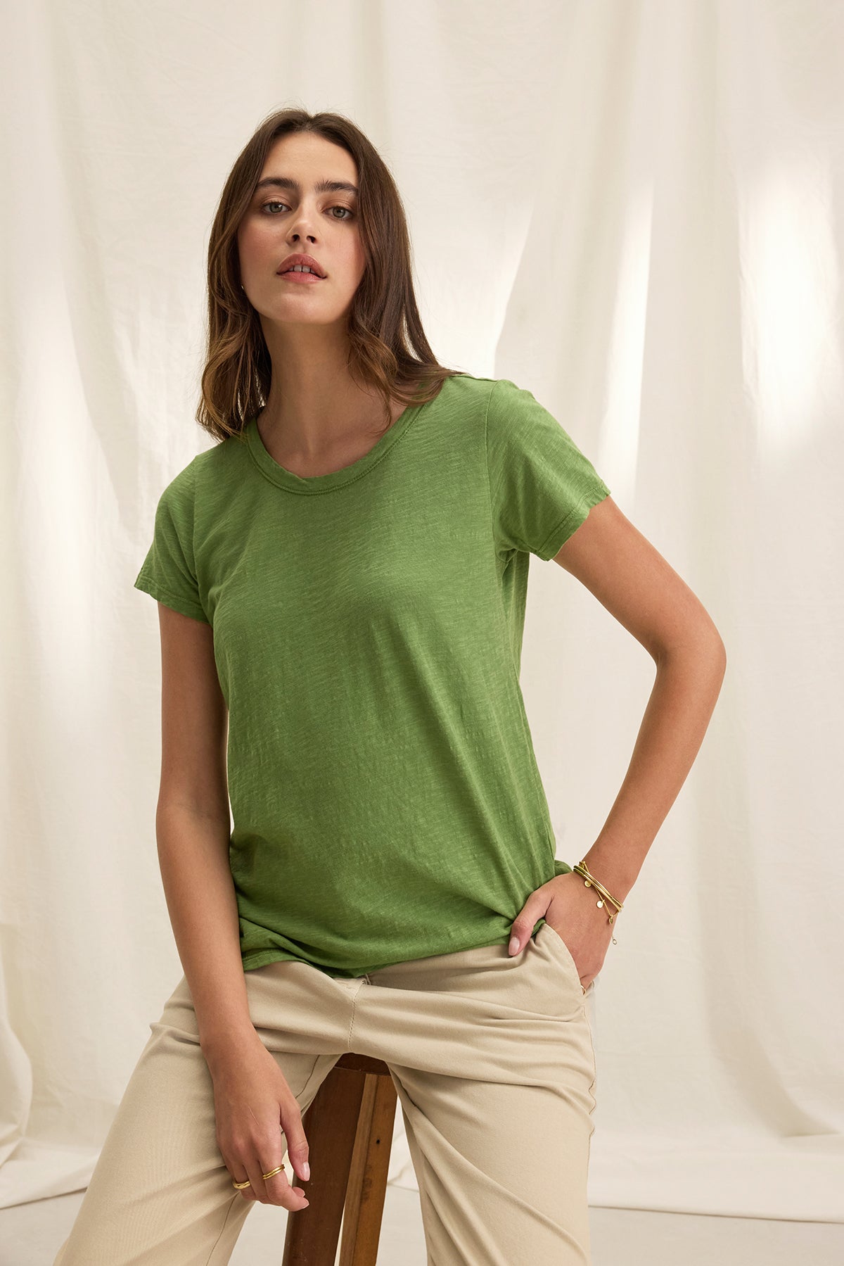   A woman in a Velvet by Graham & Spencer TILLY TEE and beige pants sits on a wooden stool against a light fabric background. 