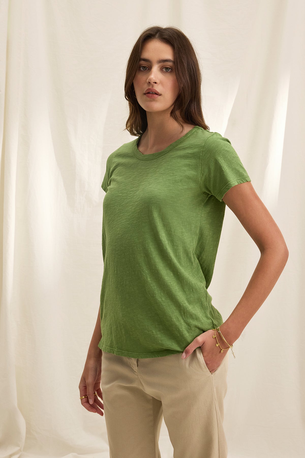 A woman stands against a beige backdrop wearing Velvet by Graham & Spencer's TILLY TEE in green and beige pants, embodying the easy-going charm of a California classic, with her hands by her sides.-38663170490561