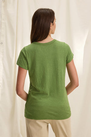 A person stands with their back to the camera against a neutral curtain backdrop, showcasing a classic California look in Velvet by Graham & Spencer's TILLY TEE in green cotton slub and beige pants.