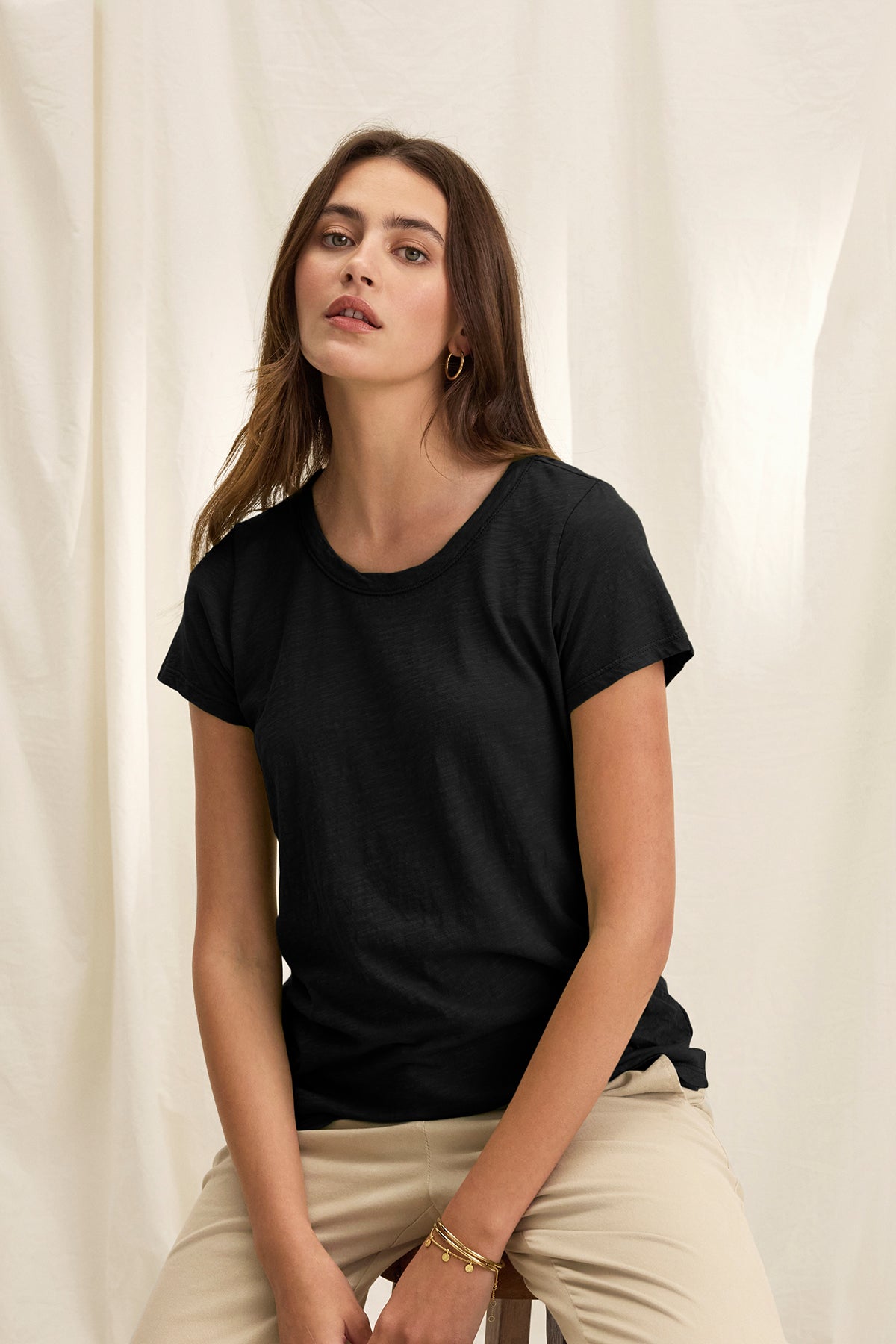   A person sits against a light fabric backdrop, wearing a TILLY TEE by Velvet by Graham & Spencer and beige pants, exuding the relaxed vibe of a California classic with their neutral expression. 