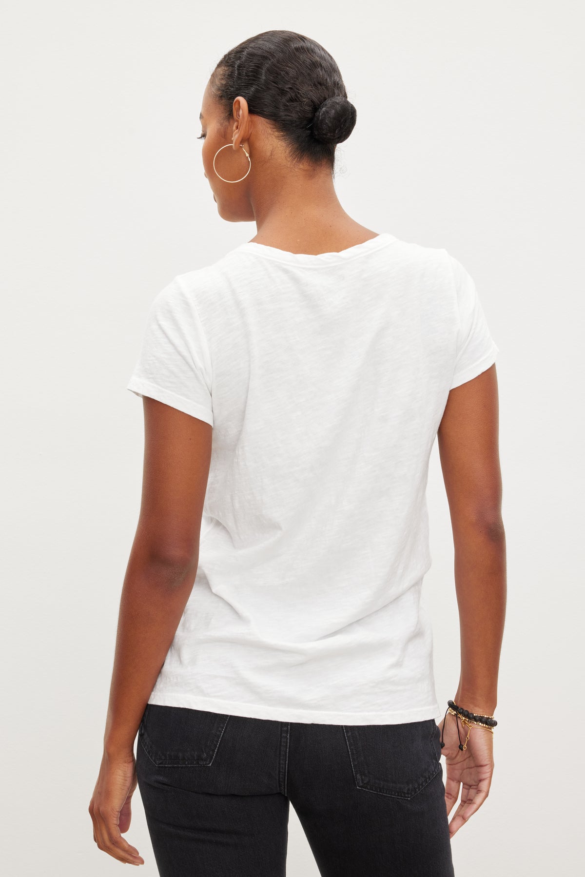   Person with hair in a bun, wearing hoop earrings and black pants, is standing facing away from the camera, showcasing a TILLY TEE by Velvet by Graham & Spencer. 