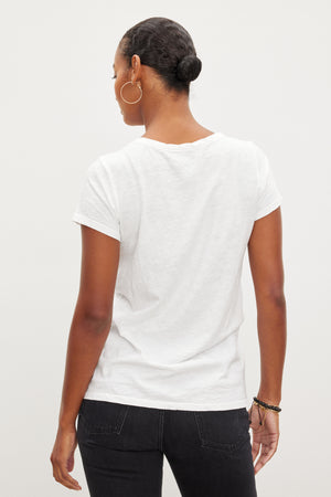 Person with hair in a bun, wearing hoop earrings and black pants, is standing facing away from the camera, showcasing a TILLY TEE by Velvet by Graham & Spencer.