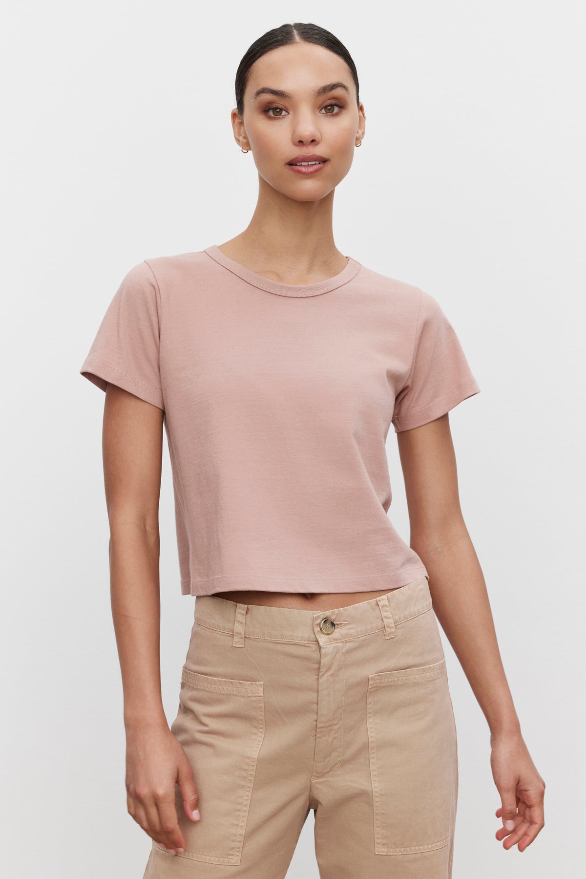   A person is standing and wearing the TINSLEY TEE, a light pink cropped tee in soft cotton by Velvet by Graham & Spencer, paired with beige high-waisted pants. They are looking at the camera against a plain white background, exuding a vintage feel. 